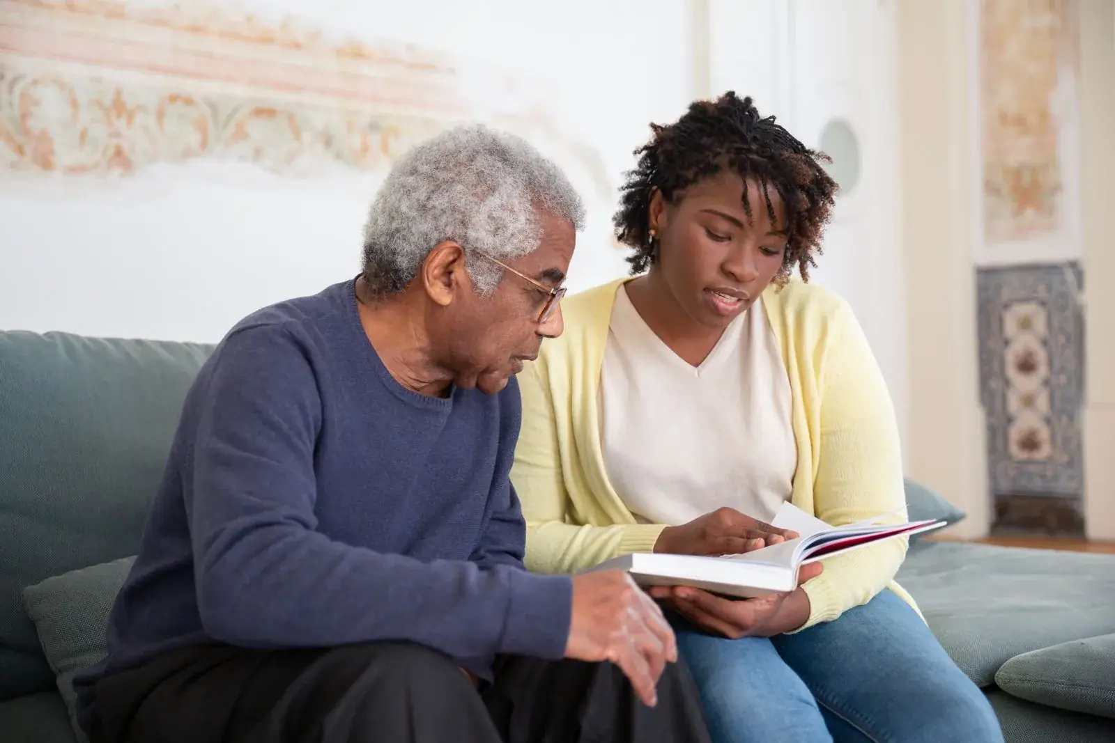 A carer reading with an elderly man on his sofa